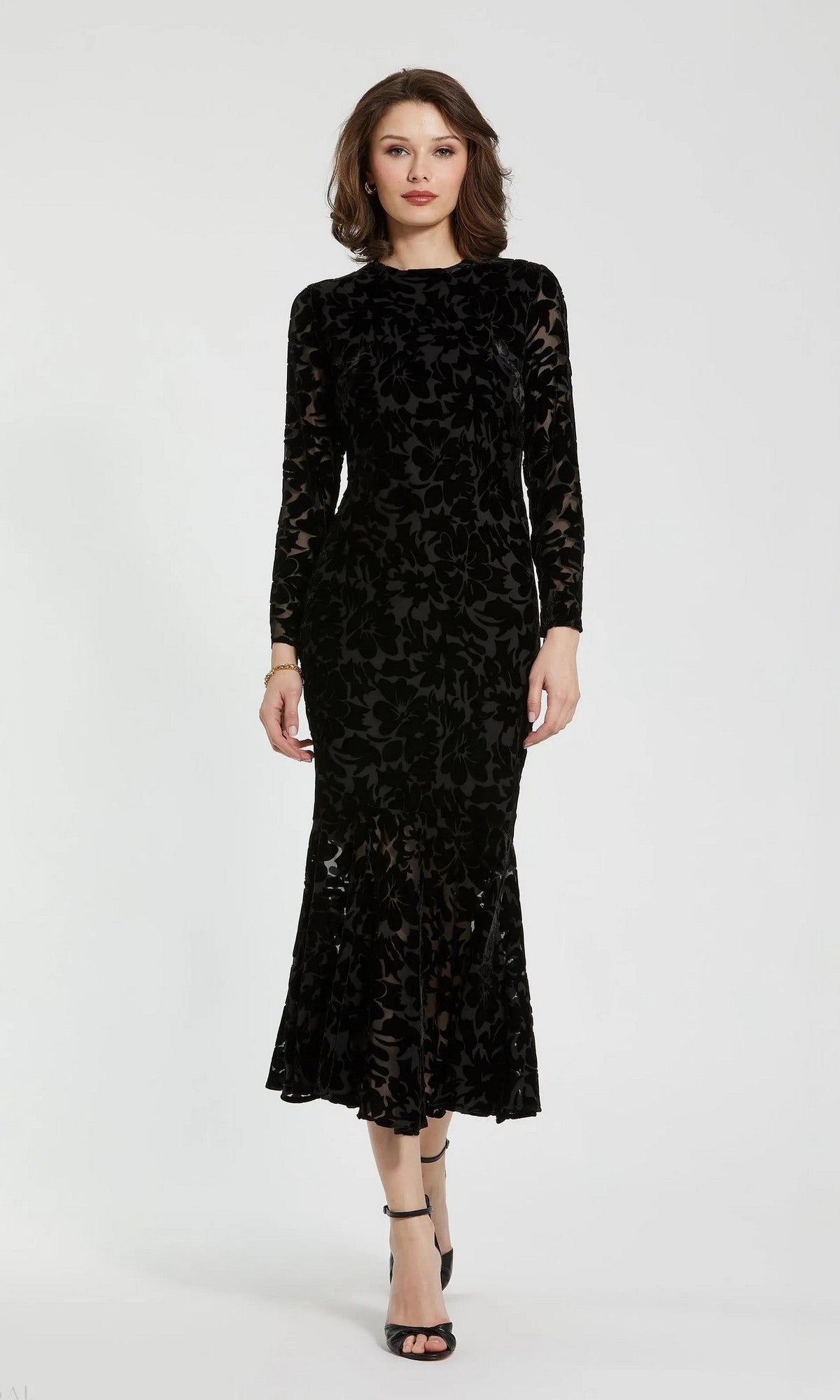A woman stands against a plain background, wearing the Mac Duggal 81129 midi party dress in black, featuring burnout velvet fabric with long sleeves and floral patterns, styled chicly with black heeled sandals.